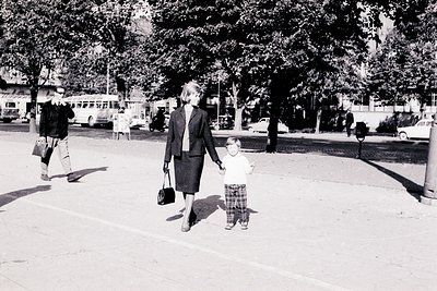 A woman in a dark suit and briefcase walks alongside a young boy in plaid shorts and sandals. A man in a trench coat walks pa...