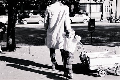 A man in a long coat walks away from the camera, pulling a young child with a distinctive wheeled toy. 1960s/70s attire: wide...