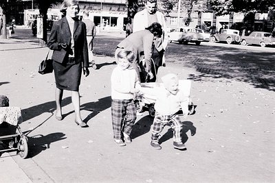 A mother and father guide two young children playing with a vintage wooden pull toy on a city street. Cars from the 1950s are...