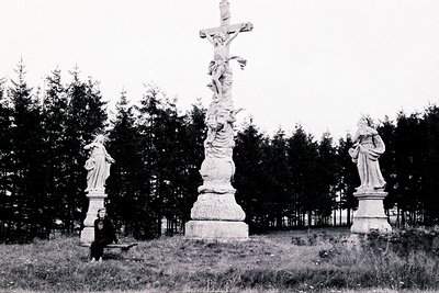 A monochrome photograph depicts a roadside shrine with a central crucifix flanked by statues of the Virgin Mary. A seated fig...