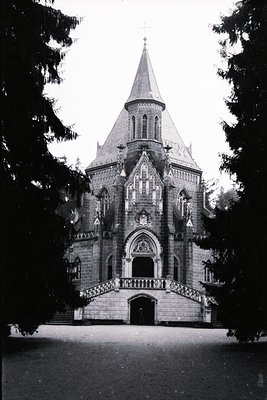 Ornate stone chapel with a pointed steeple, framed by tall evergreen trees. Features a grand staircase leading to an elaborat...
