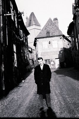 A man in a dark overcoat stands center in a narrow, cobbled street lined with historic buildings. Above, a distinctive roofli...