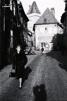 A woman in a fur-trimmed coat and heels strides down a narrow, cobbled street. The architecture suggests a European town, pos...