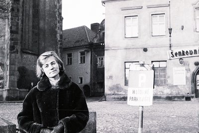 Young woman in a dark fur coat poses near a stone wall and historic building. Cobblestone street and a parking sign are visib...
