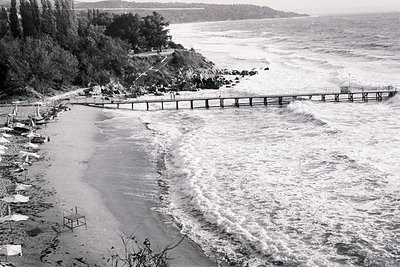 Dramatic waves crash along a pier extending into the Black Sea. A sandy beach lined with umbrellas and sparse figures stretch...