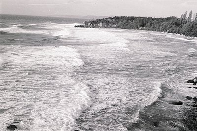 Dramatic coastal scene captures turbulent waves crashing against a rocky shoreline. Dense vegetation lines the coast, contras...