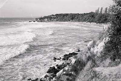 Rocky coastline with crashing waves and a sandy beach backed by dense vegetation and a distant cliff. A sailboat is visible o...