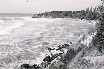 Dramatic, black and white aerial view of a rocky coastline. Waves crash against the shore, revealing a sandy beach beyond. Lu...