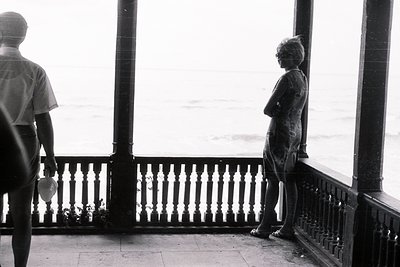 A woman stands with arms crossed, gazing out at a hazy seascape from a balcony. A young boy, holding a bottle, stands nearby....