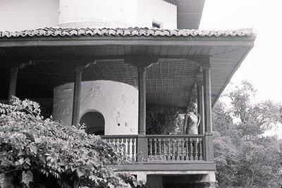 Stone balcony features a woman in profile, overlooking lush foliage. The structure shows architectural details with a rounded...