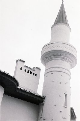 Low-angle view of a white minaret with tiered windows, typical of Ottoman architecture. Textured stucco facade and dark roofl...