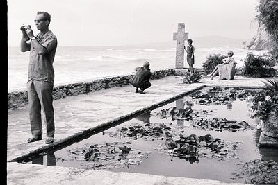 A man, likely a visitor, photographs a lily pond and coastal landscape. A large stone cross and seated figures are visible be...