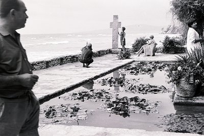 Black and white image depicting a lush, tiered garden overlooking the sea. A man stands in profile at left, a cross stands ce...