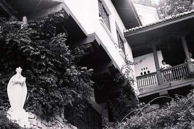 Statue of the Virgin Mary stands near a traditional building with a cantilevered balcony and whitewashed walls, heavily drape...