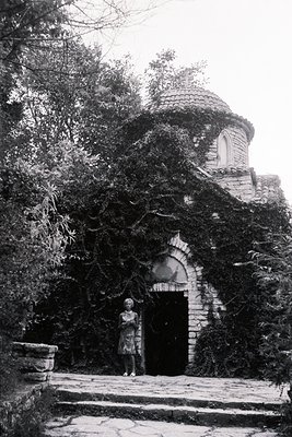 A woman stands before an ornate, ivy-covered stone building with a distinctive domed roof. The architecture hints at Mediterr...