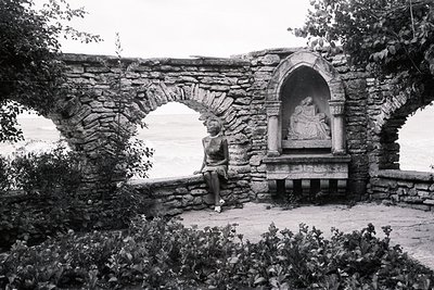 Woman sits thoughtfully within a stone courtyard, framed by arched stonework and a niche containing a statuary of Madonna and...