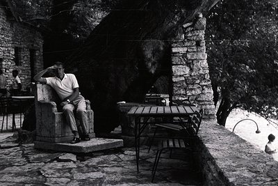 Man relaxes in a stone chair on a patterned patio. Stone walls and a partial view of foliage create a secluded, courtyard fee...