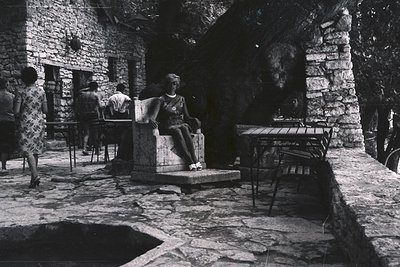 A seated woman in a patterned dress rests on a stone bench within a courtyard, framed by rough-hewn stone walls. Patrons are ...