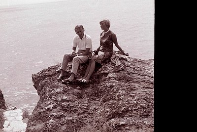 A formally dressed couple sits on a rocky outcrop overlooking a vast seascape. Man in a light-colored suit and woman in a fit...