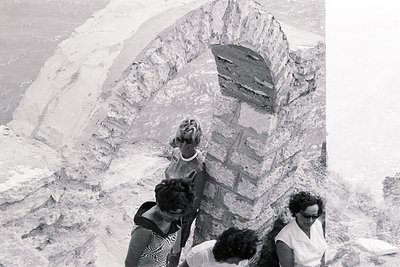 Three women in 1960s attire stand near a crumbling stone archway, likely part of a larger structure. The arch features rough ...