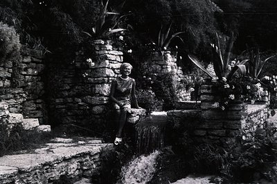 A young woman sits on a stone ledge overlooking a tiered fountain in a lush, terraced garden. Stone walls with inset planting...