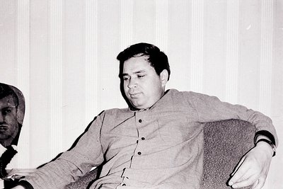 A portrait of a man seated in a plush armchair, posed in a relaxed manner. The subject wears a collared shirt and appears to ...