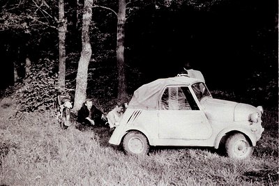 A vintage Fiat 500 convertible sits beside a grassy path, two figures appearing casually posed nearby. The car's canvas top i...