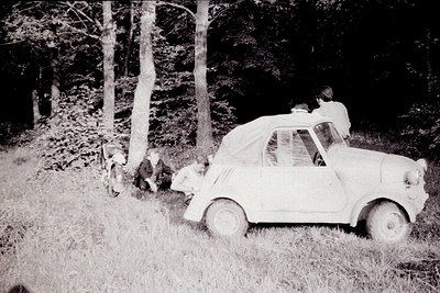 Vintage photograph depicting a small, open-top car (likely an Isetta) stranded in a grassy, wooded area. Four figures are pre...