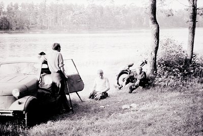 Three men beside a vintage convertible car and motorcycle, overlooking a lake. The car, likely a late 1950s or early 1960s mo...