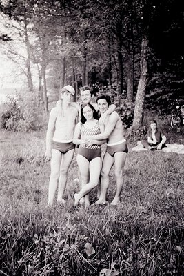 Group portrait of four figures in swimwear amidst tall grasses and dense trees, possibly near a lake or seaside. Three men fl...