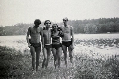 Four young adults pose by a lakeside, dressed in swimwear; indicative of leisure time. Men sport shorter, 1970s-style haircut...