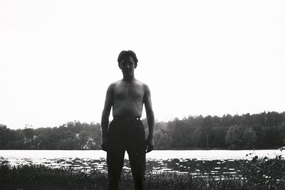 Young man stands on a grassy bank, partially clothed, facing the viewer. A tranquil lake and dense woodland backdrop create a...