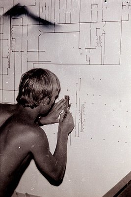 A young man, shirtless and with short hair, examines a large technical diagram with a magnifying glass. The schematic, likely...