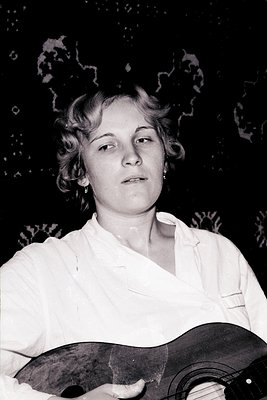 Portrait of a woman, likely a musician, holding an acoustic guitar. She wears a collared shirt, with carefully styled, curled...
