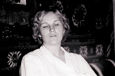 Young woman with short, curled hair, eyes closed, appears contemplative. She wears a collared shirt. Background features a ri...