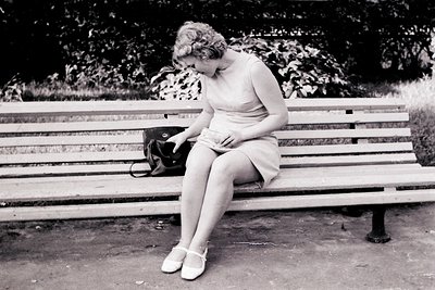 A young woman in a 1960s-style shift dress and heels sits on a wooden park bench, appearing preoccupied while examining a sma...