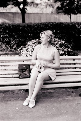 A woman in a fitted dress and heels sits on a park bench, her gaze directed off-camera. She holds a clutch purse, a handbag r...