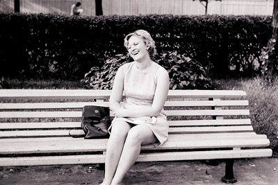A young woman, smiling broadly, sits on a park bench in a sleeveless dress. A black bag rests on the bench beside her. The sc...