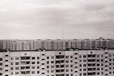 Massive, repetitive brutalist apartment block dominates the frame. Numerous windows and arched balconies punctuate the stark ...