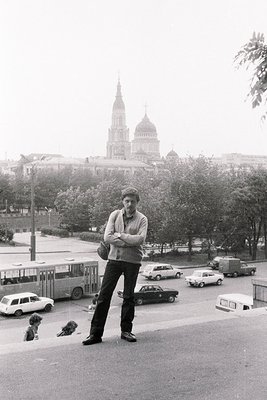 A man with short hair stands arms crossed, facing the camera, with a tall church dome visible in the background. Appears to b...