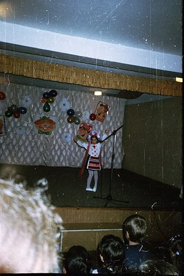 A performer in traditional folk attire balances juggling pins on a string, surrounded by suspended fruit and vegetables in a ...
