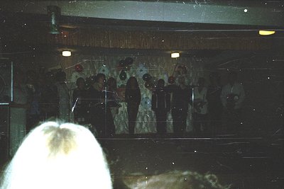 Vintage indoor performance featuring a choir or band in formal attire, likely 1970s–1980s. Stage backdrop includes a mural wi...