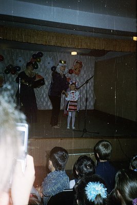 A staged puppet performance in a dimly lit indoor venue, likely a school or community hall. Two puppeteers manipulate charact...