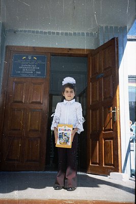 Young student in traditional Ukrainian school attire (white blouse, dark skirt, white headscarf) stands outside a Soviet-era ...