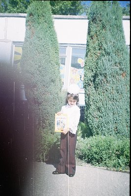 Young girl in 1970s-style dress and skirt stands outdoors holding a vintage book, likely a children’s illustrated novel. Gree...