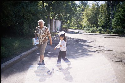 Vintage black-and-white street scene: elderly woman in patterned dress and sandals holds a book, guiding a young girl in a wh...