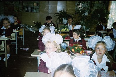 Vintage school cafeteria scene with students in early 1970s Soviet-style uniforms—white blouses, red vests, and large white h...