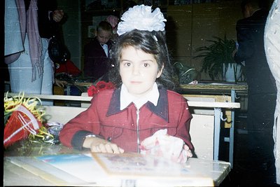 Young girl in a red school uniform with a white bow hair accessory, seated at a wooden desk in an indoor classroom setting. T...