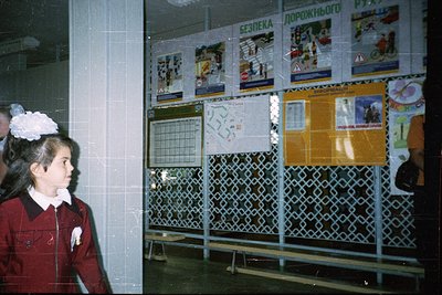 Soviet-era school hallway with traffic safety posters in Cyrillic (#БезопасностьДорожногоДвижения). Girl in maroon school uni...