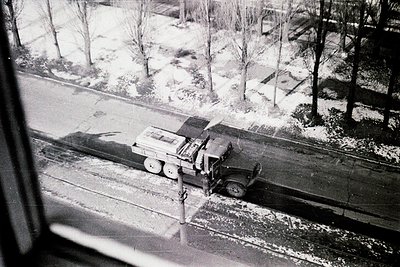 Vintage black-and-white photo of a vintage military truck navigating snow-covered urban streets, likely mid-20th century. Sno...